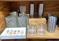 Wide view showing two glass decanters with diamond pattern; two glass tumblers; four cylindrical stainless steel cups; wooden cigar box; box with five chess piece bottle stoppers.