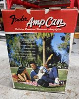 Box front showing Fender Amp Can battery powered portable amplifier, key features and lifestyle photo with people playing guitar outdoors.