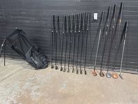 Full view of 11 left handed iron clubs, 3 wood clubs, and 1 putter lined up beside black Ping golf stand bag on a concrete floor against black wall.