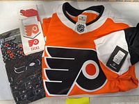 Photo showing orange and white Philadelphia Flyers Fanatics jersey with NHL shield, socks with Flyers logo in packaging, and adjustable face mask with Flyers logos in packaging.