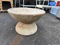 Wide side view of heavy concrete planter with pedestal base on asphalt surface.