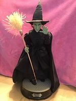 Photo of the 19-inch tall Wicked Witch of the West figurine from the Wizard of Oz with black cloth cape, hat, green face, and broomstick on a circular base.