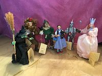 Photo of 6 Wizard of Oz themed FABRICHE collectable figurines arranged side by side: Witch, Cowardly Lion, Scarecrow, Dorothy, Tin Man, and Glinda, each with tags attached.