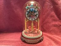 Front view of the Wizard of Oz music box clock with glass dome, featuring the clock face with the characters, gold pillars, rotating red bird pendulum, and decorative base.