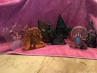 Full set of six colored clear resin Wizard of Oz character figurines on pink towel: Glinda, Scarecrow, Tin Man, Cowardly Lion, Dorothy with Toto, Wicked Witch of the West.
