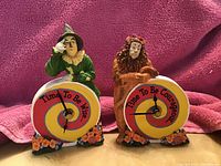 Photo showing two Wizard of Oz themed clocks featuring Scarecrow and Cowardly Lion figurines with colorful clock faces bearing motivational text.