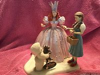 Photo showing three Wizard of Oz-themed ceramic snow baby figurines: Glinda, Dorothy, and Toto, arranged on a snowy white base against a pink background.