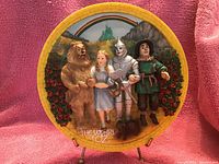 Photo of a decorative round plate with painted characters from The Wizard of Oz including Lion, Dorothy, Tin Man, and Scarecrow with rainbow and castle background.