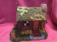 Front and side view of a small hand-painted ceramic or resin cottage figurine with green textured roof and floral decorations representing the House of Tin Man from the Wizard of Oz collectible series.