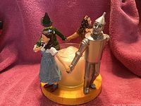 Four Wizard of Oz characters arranged in a circle holding hands around a frosted glass candle holder on yellow round base, displayed against a pink fabric background.