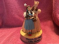 Photo of two Wizard of Oz figurines on pink fabric backdrop showing Dorothy and Glenda standing on base reading 'There's No Place Like Home'