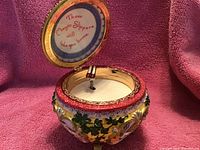 Open view of ornate round music box with floral design and red glittery rim, showing inscription on inner lid, and rotating ruby slippers figurine inside.
