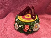 Music box with ruby red slippers on display against a pink textured cloth background. Detailed close-up shows floral decoration around the base and yellow platform with swirl pattern.