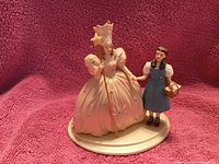 Photo shows two porcelain figurines on a round base. Glinda the Good Witch in white gown with a crown and wand, and Dorothy in blue dress holding a basket. Photo is taken to highlight the ornament details.