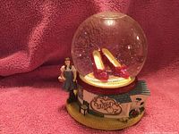 Snow globe with a clear glass sphere containing ruby slippers and a Dorothy figurine attached to the base, displayed against a red fabric background.