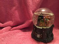 Photo showing a Wizard of Oz themed snow globe with Wicked Witch and Flying Monkey figurines inside clear glass dome. The base resembles a castle with barred windows and textured stone effect.