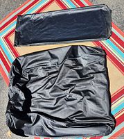 Black cartop carrier laid out partially folded on outdoor fabric mat. Fabric has faint wrinkling and some folding marks.