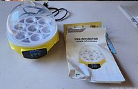 Yellow and white egg incubator with transparent cover and user manual placed beside it on table.