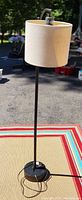 Full view of the black metal floor lamp with a beige linen drum shade standing on a colorful mat outdoors, showing cord and base.