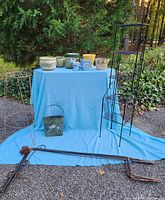 Full view of garden items on blue fabric outdoors including ceramic pots, plant stands, metal poles, metal pocket planter, and flag holder.