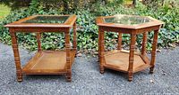 Two wood and glass top end tables with rattan base, one rectangular and one hexagonal, placed outdoors on a paved surface with greenery in the background.