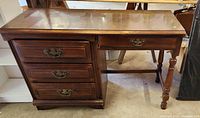 Front-side view of vintage wooden desk showing three drawers, turned wooden leg, and worn desktop surface.