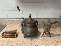 Front and side view of the three items together on countertop: metal oil lamp with tripod legs and two handles, metal fire starter tool inserted into lamp lid, and vintage box of Maple Leaf matches.