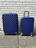 Full view of two Heys luggage pieces on pavement against brick wall, showing size difference and details like wheels and handles.