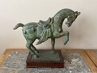 Front and side views of an iron horse sculpture mounted on a wooden base, showing the green patina and detailed saddle design.
