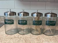 Front view of all four vintage glass containers with metal lids and green labels for bandages, cotton, tongue depressors, and applicators.