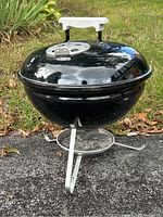 Front view of the black portable Weber charcoal grill showing the lid with white handle and vent on top, standing on a silver tripod stand outdoors.