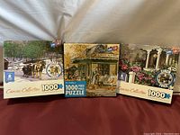 Three unopened 1000 piece puzzle boxes showing various scenic and vintage house themes.