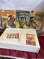 Photo showing three vintage comics: Zane Grey's Wilderness Trek, Walt Disney Sword and the Rose, Walt Disney Wringle Wrangle, and the Canadian Superheroes stamp book partially visible on a box.