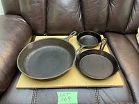 Three cast iron pans: largest pan is WagnerWare 11 3/4", medium pan W.K.M 8", and smallest 6.5". Positioned on cardboard on brown leather seating, each pan has a handle with a hanging hole, pouring spouts on sides. Some surface marks and general wear visible.