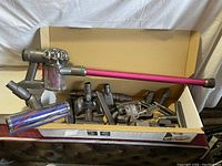 Dyson V6 Animal handheld vacuum with purple extension wand laid inside a cardboard box with multiple accessories and parts.
