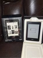 Kindle Paperwhite e-reader device inside original box with packaging visible. Device screen shows low battery icon on e-ink display.