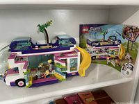 Photo of assembled Lego Friends 41395 Friendship Bus showing the two-level structure, yellow slide, open roof with tree, minifigures inside, and some loose pieces in bag.