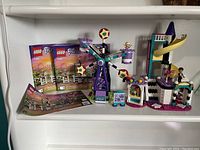Lego Friends set 41689 with Ferris wheel, slide structure, minifigures, and accessories on a white shelf with three instruction manuals visible.