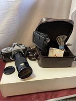 View of Nikon Nikomat camera with attached zoom lens, camera case opened showing various accessories inside including cables and Hoya filter box.