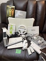 Full setup of Nintendo Wii console, balance board, game, multiple controllers, charging station, steering wheel accessory, manuals, and cables on a chair.