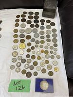Top-down view of approximately 70 old coins arranged on fabric, showing variety of sizes, metals, and designs. Metal storage box and single displayed coin in blue holder included.