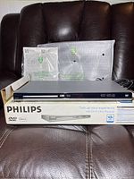 Photo of the silver Philips DIVX Ultra DVD Player sitting on its original box, with manuals and cables behind it on a brown leather couch.