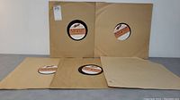 Five 16 inch records in plain brown paper sleeves arranged in a semicircle on a flat surface.