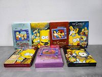 Front view of seven The Simpsons complete season DVD boxed sets arranged in two rows, showing colorful character artwork on covers.