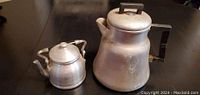 Front view of the small aluminum teapot next to the larger aluminum coffee pot on a dark surface.