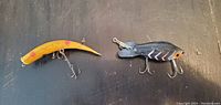Photo showing two fishing lures placed on a dark surface, one yellow curved lure with red marks and three silver treble hooks, and one black fish-shaped lure with orange eyes and a metal diving lip, both with three treble hooks.