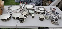 Photo of ceramic dinnerware set arranged on white folding tables outside, showing plates, bowls, mugs, and condiment dishes with blue and beige decorative pattern.