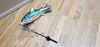Fishing pole with a metallic fish-shaped lure attached, showing the lure details and coiled wire mechanism on wooden floor.
