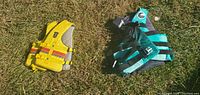 Two adult life jackets, one yellow with reflective strips and one green with black straps, laying on grass.