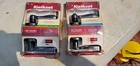Two Kwikset door handles in original packaging placed side-by-side on a surface, showing front of packages with product and branding details